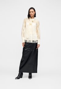 Δαντελένια μπλούζα σε κρεμ χρώμα με δετή λαιμόκοψη, διάφανα μακριά μανίκια· συνδυασμένη με μαύρη δερμάτινη φούστα μάξι και μαύρες μπότες με τακούνι και μεταλλικές λεπτομέρειες.