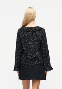 Γυναίκα με μακριά ξανθά μαλλιά, που φοράει μαύρο πουκάμισο με πουά και βολάν στο γιακά και στις μανσέτες, καθώς και μαύρη φούστα, γυρισμένη πλάτη σε λευκό φόντο.