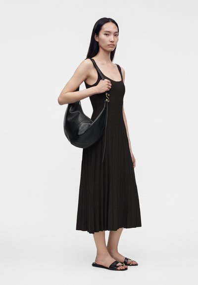 Femme aux longs cheveux noirs portant une robe midi noire plissée sans manches, des sandales plates noires, et portant un grand sac à bandoulière en cuir noir.