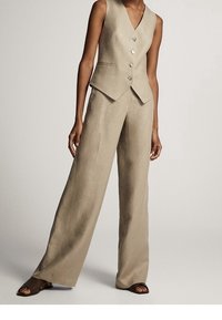 Donna con un gilet smanicato beige con bottoni e pantaloni wide-leg abbinati a sandali aperti marroni, in piedi su uno sfondo neutro.