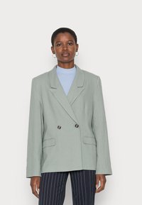 Blazer de dupla face em tecido verde claro, com lapelas de bico, dois bolsos frontais e botões escuros. Usado sobre um suéter de gola alta azul.