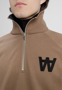 Brązowy pulower na suwak z wysokim kołnierzem, ozdobiony czarnym haftowanym logo i gładką fakturą. Widoczne zapięcie na suwak.