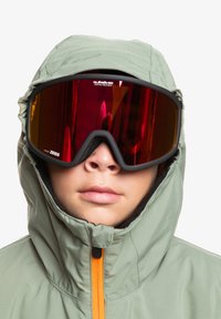 Lyžařské brýle s velkým černým rámem a červenými zrcadlovými skly. Nosené s zelenou bundou s kapucí, která má oranžový zip.
