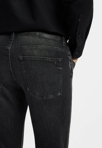 Czarne jeansy z denimu z wyblakłym wykończeniem, o konstrukcji z pięcioma kieszeniami i oznakowaną łatą z tyłu. Widoczne są przeszycia oraz gładka tekstura.