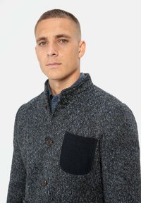 Grijze gebreide blazer met een textuurpatroon, knopen aan de voorkant en een donkerblauwe opgestikte zak. Staande kraag en aansluitende pasvorm.