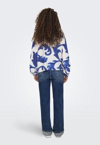Blusa bianca con motivo floreale blu, vestibilità ampia e maniche lunghe, abbinata a jeans dritti blu scuro con risvolti.