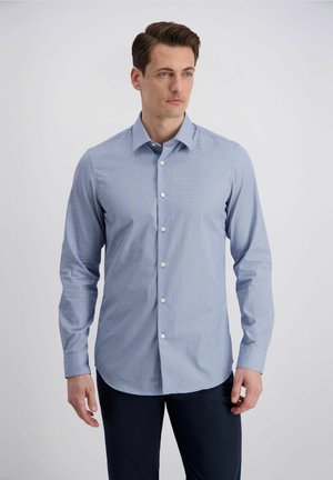 Mann trägt hellblau gemustertes, langärmeliges Hemd und dunkle Hose, steht vor schlichten hellem Hintergrund und schaut nach rechts.