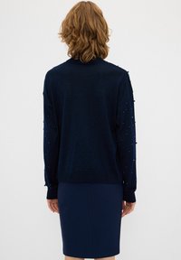 Marineblau gestrickter Pullover mit strukturierten Punktakzenten an den Ärmeln, gerippten Bündchen und Saum. Kombiniert mit einem schlichten marineblauen Bleistiftrock.