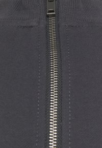 Gros plan sur un tissu noir avec une fermeture éclair en métal argenté centrée, mettant en valeur les détails des coutures de chaque côté de la fermeture éclair.