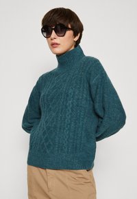 Türkiis turtleneck särk, millel on tekstuuriga teemantmuster ja ribsukantidega varrukad. Üks suurus suurem. Sobitatud suurte ümmarguste päikeseprillidega.