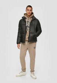 Schwarze Pufferjacke mit Kapuze, ausgestattet mit einem Front-Reißverschluss und gesticktem Logo, über einem beigen Sweatshirt und passenden Jogginghosen getragen.