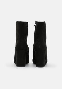 Bottines en daim noirs avec une finition lisse, talons bloc hauts et fermetures éclair sur le côté. Design minimaliste sans quincaillerie visible.