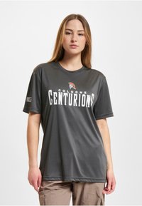 Tamsiai pilkas trumpomis rankovėmis marškinėliai, pagaminti iš lygaus audinio, su užrašu "COLOGNE CENTURIONS" baltu šriftu ir spartiečių logotipu viršuje.
