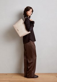 Beige kožená kabelka typu tote s strukturovaným designem, která má dva akcenty ve zlatém tónu a hladký povrch. Modelka má na sobě černý top a hnědé kalhoty.