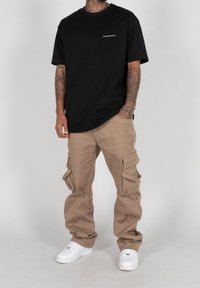 Maglietta nera in cotone con maniche corte e un piccolo logo bianco sul petto, abbinata a pantaloni cargo beige con più tasche laterali. Sneakers bianche.