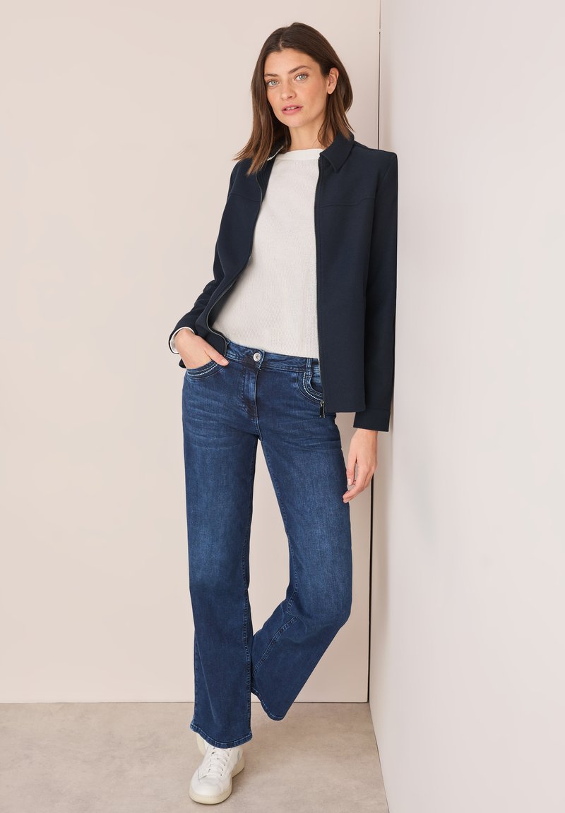 Maritieme blazer, beige trui en blauwe flare jeans. Witte sneakers met gestructureerde zolen. Gladweefsel en strakke lijnen. Minimalistisch ontwerp.