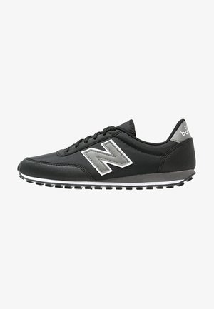 Czarny sneaker New Balance z szarym logo, białym paskiem na podeszwie, wiązaniem na sznurówki i teksturowaną gumową podeszwą, widziany z boku.