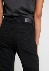 Černé denimové džíny s vysokým pasem, se koženou nášivkou a dvěma zadními kapsami, přičemž jedna je ozdobena malým červeným, bílým a modrým logem.