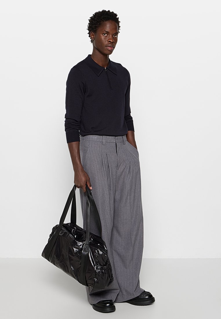 Polo negro de punto con cuello, pantalones de pierna ancha gris a rayas, y un bolso de viaje negro brillante. Textura suave, ajuste casual.