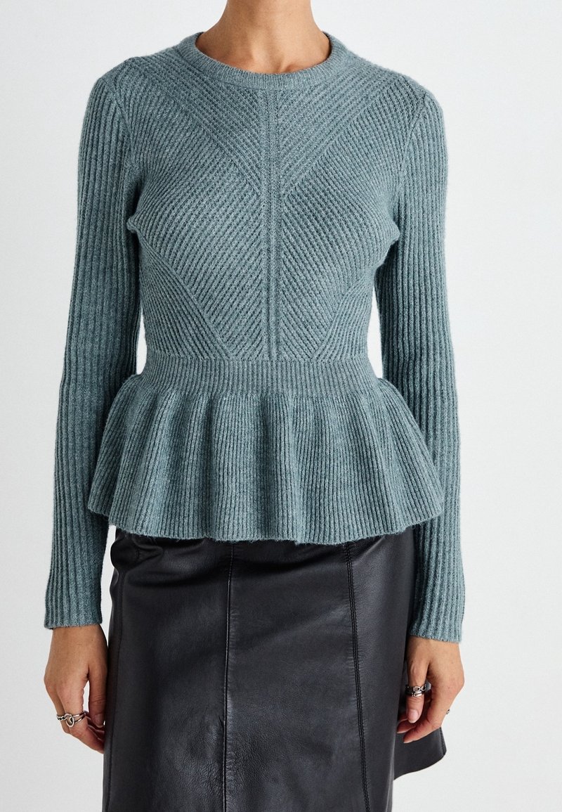Pull en tricot côtelé à manches longues couleur teal avec taille peplum associé à une jupe en cuir noire, porté par une personne avec des bagues aux doigts.