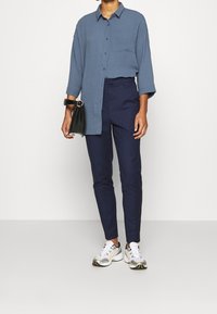 Chemise bleue ample à boutons avec une poche poitrine, associée à un pantalon bleu marine ajusté et des baskets blanches avec des accents argentés et jaunes.