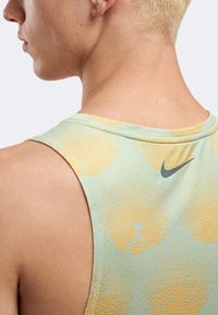 Κοντινό πλάνο ενός ατόμου με κοντά ξανθά μαλλιά, που φοράει πράσινο αμάνικο τοπ με πορτοκαλί πουά και ένα μικρό γκρι λογότυπο της Nike στην πλάτη.