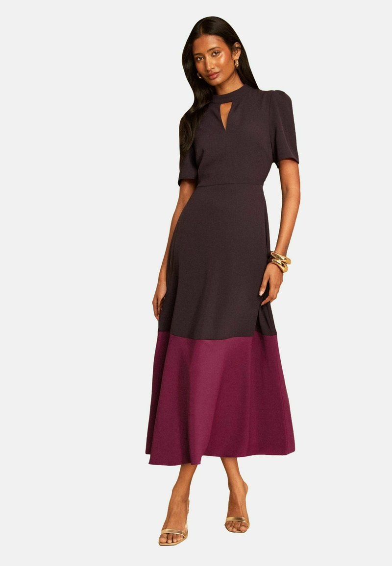 Woman wearing a black and purple short-sleeve midi dress with keyhole neckline and gold sandals, standing against a plain background.