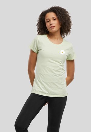 Femme aux cheveux bouclés portant un t-shirt vert clair avec une petite marguerite blanche sur la poitrine et un pantalon noir sur un fond gris.