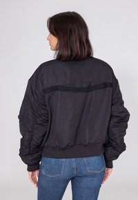 Czarna kurtka bomberka o luźnym kroju, z elastycznymi mankietami i dolnym brzegiem, żebrowanym kołnierzem oraz gładką, błyszczącą teksturą.