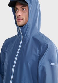 Giacca impermeabile blu con colletto alto e cappuccio regolabile. Caratterizzata da una chiusura frontale con zip e un logo bianco sulla manica destra. Texture liscia.