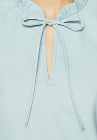 Prenda de color azul claro con un escote en forma de ojo de cerradura y un lazo atado, además de un cuello con volantes. La tela parece suave y con un ligero brillo.