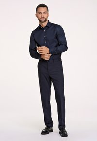 Camisa de vestir azul marino con mangas largas, con botones en el frente, combinada con pantalones oscuros y zapatos de cuero negros. Textura suave, corte ajustado.