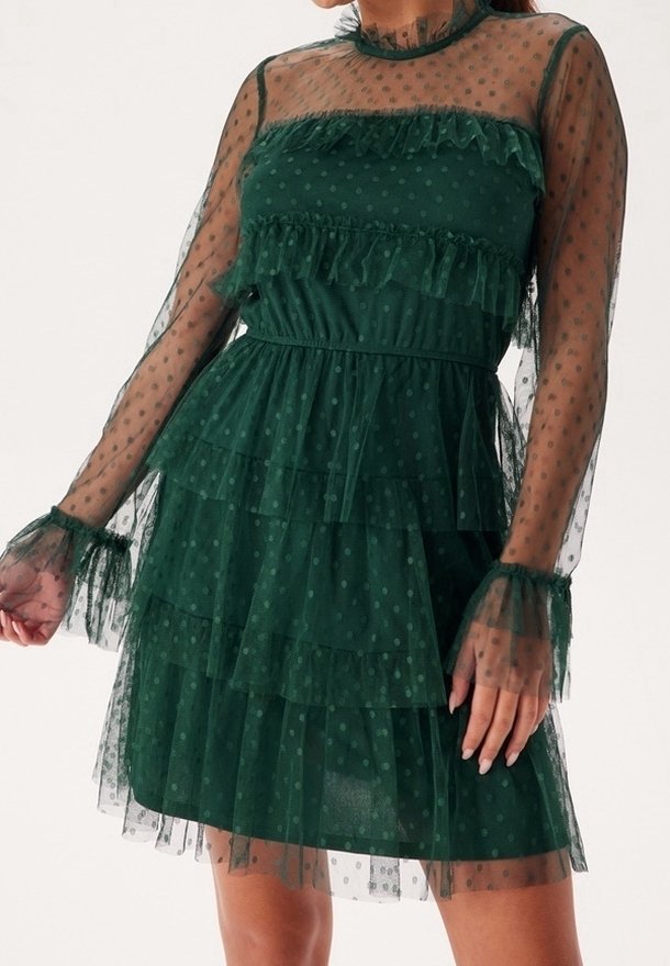 Robe verte transparente à pois avec des volants superposés, manches longues transparentes et taille cintrée portée par une personne debout.