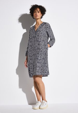 Abito navy con motivo a onde bianche, scollatura a V, maniche lunghe e tasche laterali. Indossato con sneakers bianche, su uno sfondo semplice.