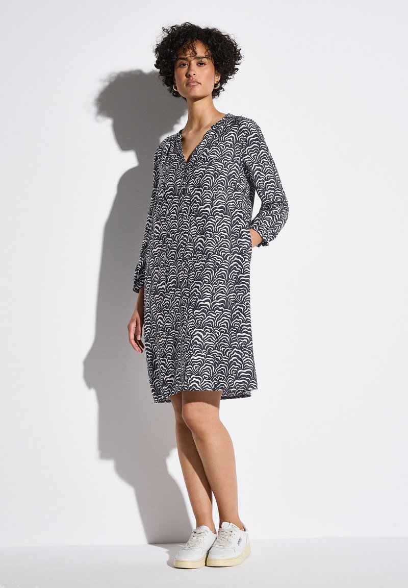 Marineblaues Kleid mit weißem Wellenmuster, V-Ausschnitt, langen Ärmeln und seitlichen Taschen. Tragen mit weißen Sneakern, vor einem schlichten Hintergrund.