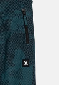 Tkanina u tirkiznoj i crnoj boji s uzorkom sadrži zip džep i brendiranu oznaku s geometrijskim logotipom, ističući funkcionalne dizajnerske elemente.