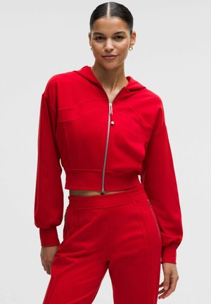Vrouw draagt een rode cropped zip-hoodie met ballonmouwen en bijpassende, hooggetailleerde broek met zijritsen, staand tegen een effen achtergrond.