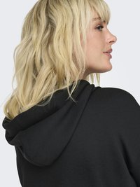 Femme blonde aux cheveux ondulés portant un sweat à capuche noir, vue de côté et de dos sur un fond clair uni.