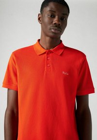 Camisa polo roja hecha de tela texturizada, con cuello y tres botones. "París" está bordado en cursiva en el lado izquierdo.