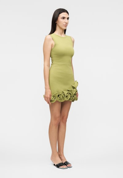 Γυναίκα στέκεται, φοράει αμάνικο ανοιχτό πράσινο φόρεμα με βολάν στο τελείωμα και μαύρα ανοιχτά, επίπεδα παπούτσια σε απλό φόντο.