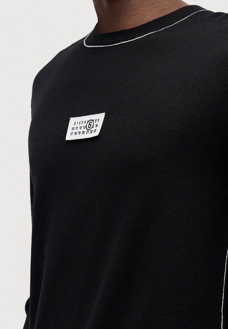 Person trägt einen schwarzen Pullover mit weißen Nähten am Kragen und einem kleinen rechteckigen Etikett mit Zahlen auf der Brust.