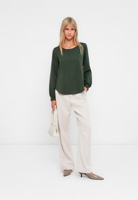 Femme portant un chemisier à manches longues vert olive, un pantalon large crème, des escarpins beige à bout pointu, tenant un petit sac à main blanc.
