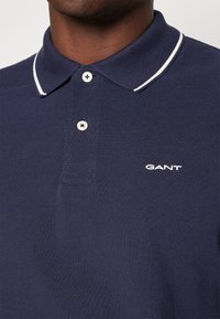 Personne à la peau foncée portant un polo GANT bleu marine avec une bordure blanche au col et deux boutons blancs fermés.