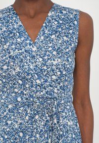 Robe sans manches avec motif floral bleu et blanc, col en V et ceinture en tissu nouée à la taille sur une personne à la peau foncée.