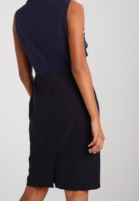 Robe sans manches bleu marine avec un design ajusté, fermeture éclair dans le dos et une fente à l'ourlet pour faciliter les mouvements. Texture lisse.