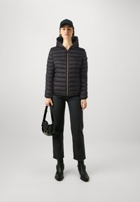 Svart dunjakke med høy krage, glidelås foran og ribbede mansjetter, sammen med mørke jeans og svarte ankelstøvler, holder en liten veske.