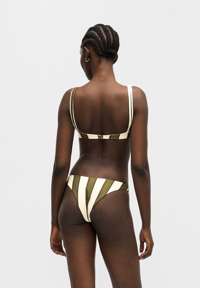 Γυναίκα με πλεξούδες που φοράει πράσινο και λευκό μπικίνι με κάθετες ρίγες, εμφανιζόμενη από πίσω σε απλό ανοιχτό φόντο.