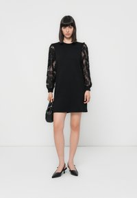 Black dress with transparent, textured sleeves adorned with floral appliqués. Model holds a small black handbag, wearing black heeled shoes.