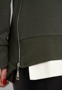 Sweat-shirt vert olive avec une fermeture éclair et un détail de tag en argent, avec un ourlet blanc contrastant en dessous et une texture de tissu lisse.