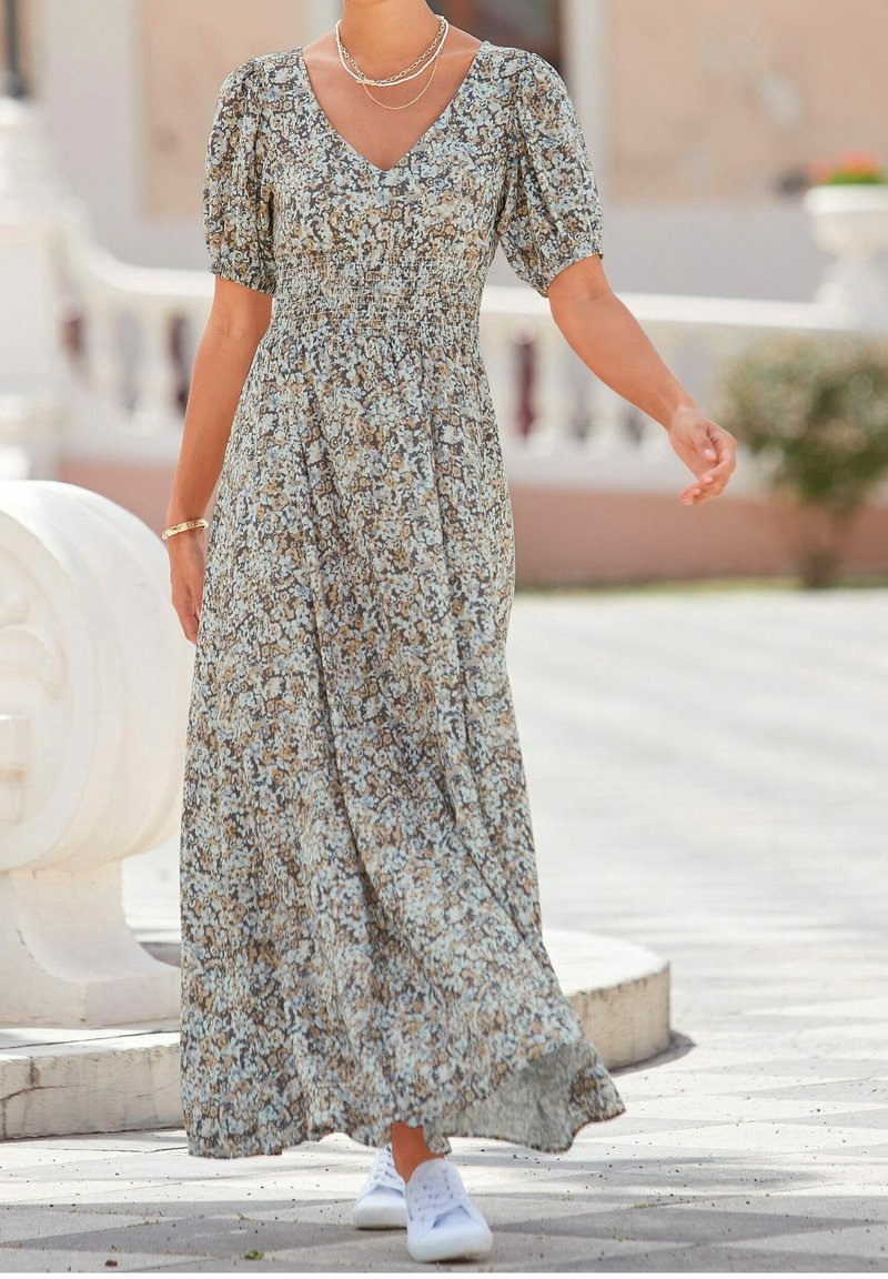 Femme marchant en extérieur portant une robe midi fleurie à manches bouffantes, des baskets blanches et des colliers en or superposés.